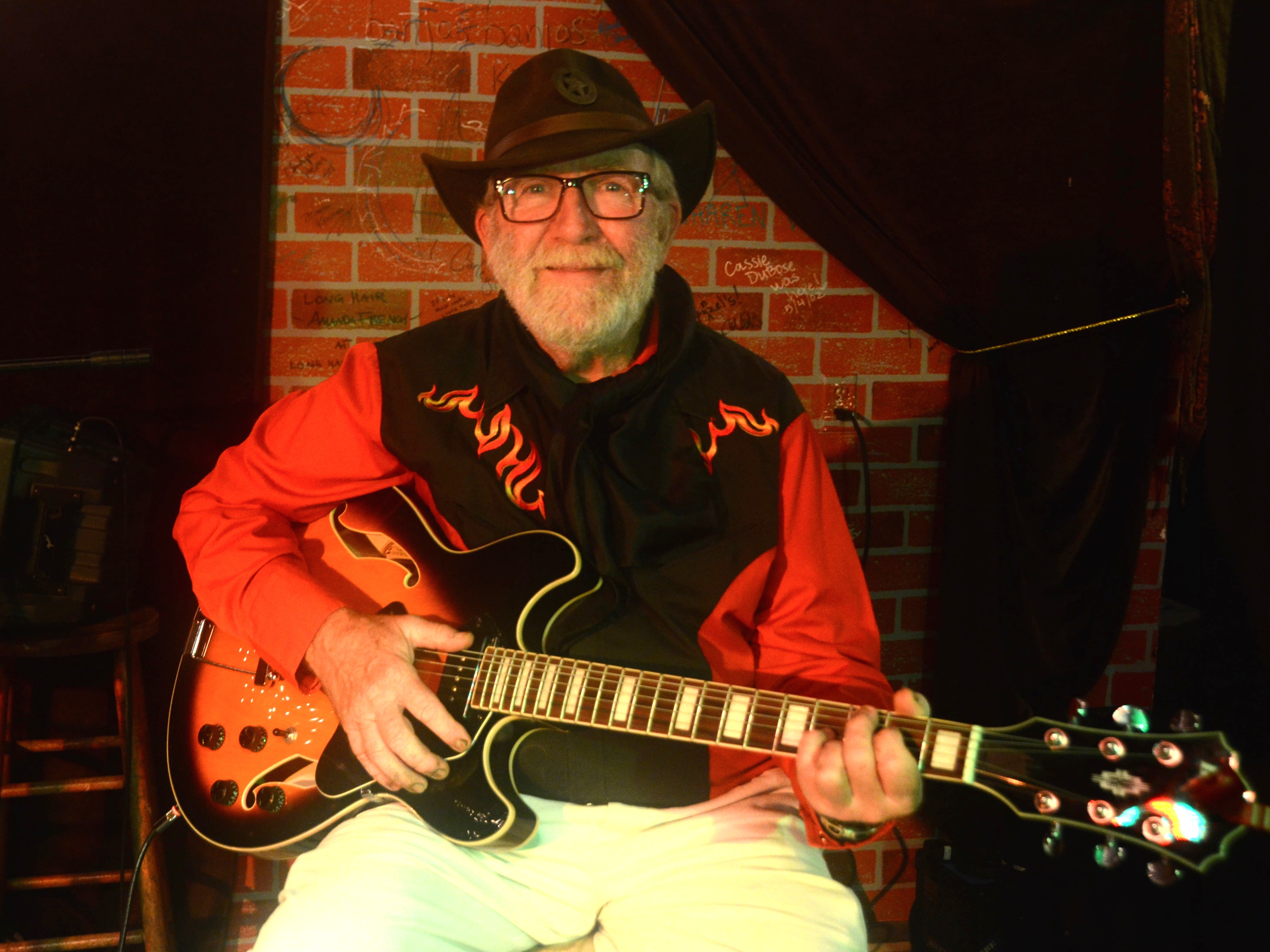 Bob Taylor seated on stage at Rapunzel&rsquo;s, playing guitar, wearing his brown cowboy hat and red shirt.