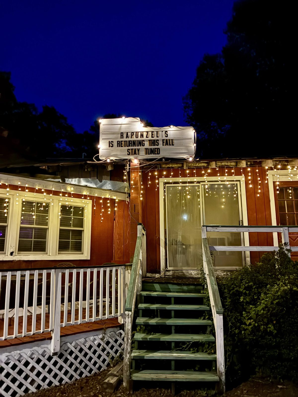 Rapunzel's at dusk: the white letterboard marquee above the barn-red building reads RAPUNZEL'S IS RETURNING THIS FALL, STAY TUNED, under strings of warm lights.